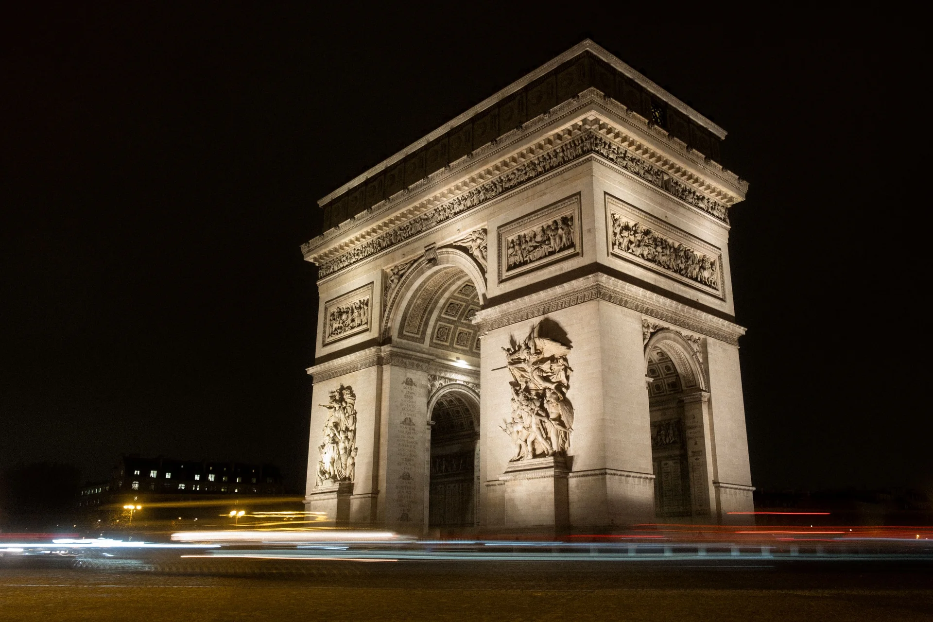 Vítězný oblouk zalitý nočním osvětlením, jeho zdobené pilíře září proti černé obloze a přes náměstí Place Charles de Gaulle se táhnou světelné stopy automobilů